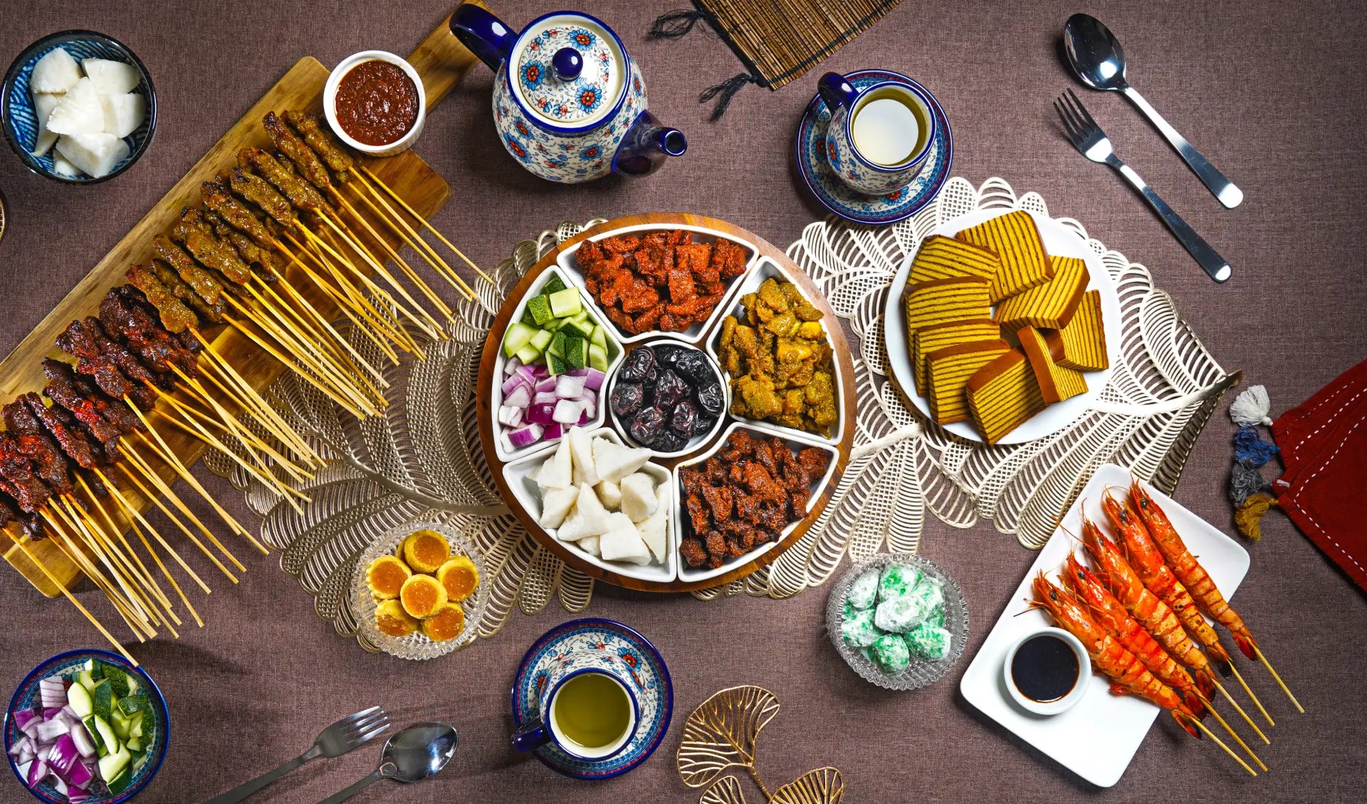 An aerial shot of a variety of Malay dishes, including skewers of satay, a platter of different curries, and slices of kueh lapis, arranged on a table.
